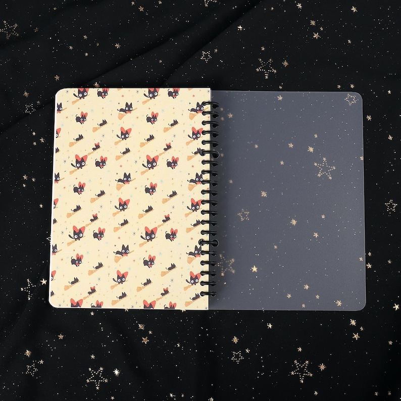 Puede incluir: Un cuaderno de espiral con una cubierta amarilla con un patr&oacute;n repetido de gatos negros montando escobas. El cuaderno tiene una cubierta de pl&aacute;stico transparente.