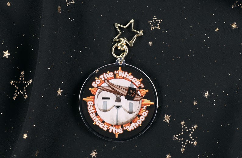 The Back of the Esquie Keychain. It features Esquie's head from Clair Obscur Expedition 33 featuring the text: "So the "Wheee" balances the "Whooo". He has the Expedition 33 Arm Band that Verso Gave him on his head.
