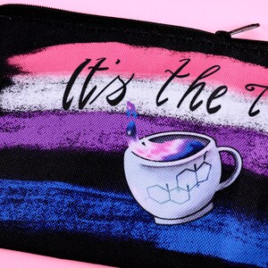 Puede incluir: Bolsa negra con cremallera con la frase "It's the T?!" y una ilustraci&oacute;n de taza de caf&eacute;. El fondo presenta pinceladas en rosa, blanco, morado y azul. La taza tiene un dise&ntilde;o de galaxia en el interior y gr&aacute;ficos de estructura qu&iacute;mica.