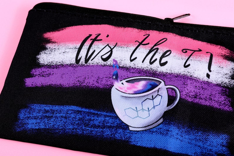 Puede incluir: Bolsa negra con cremallera con la frase "It's the T?!" y una ilustraci&oacute;n de taza de caf&eacute;. El fondo presenta pinceladas en rosa, blanco, morado y azul. La taza tiene un dise&ntilde;o de galaxia en el interior y gr&aacute;ficos de estructura qu&iacute;mica.