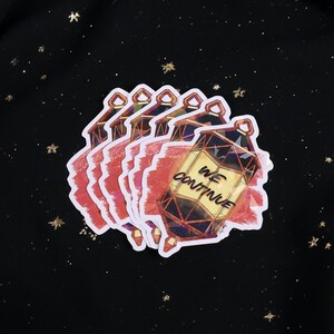 The Holographic lumina converter sticker with the text "We Continue" on top. Behind the keychain is a red paint swipe.