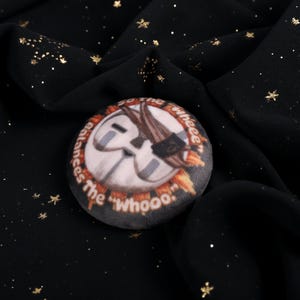 A Squishy Plush pin of Esquie from Expedition 33-  It features Esquie's head from Clair Obscur Expedition 33 featuring the text: "So the "Wheee" balances the "Whooo". He has the Expedition 33 Arm Band that Verso Gave him on his head.