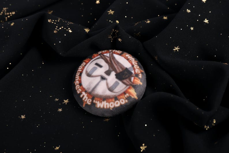 A Squishy Plush pin of Esquie from Expedition 33-  It features Esquie's head from Clair Obscur Expedition 33 featuring the text: "So the "Wheee" balances the "Whooo". He has the Expedition 33 Arm Band that Verso Gave him on his head.