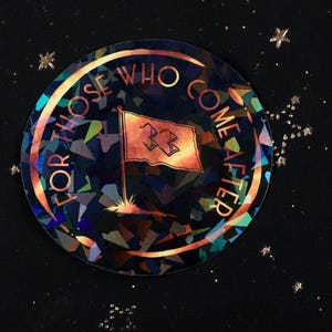 May include: A round sticker with the text "FOR THOSE WHO COME AFTER" in gold lettering. The sticker features a flag design in the center, with a geometric, iridescent background. The background is dark with gold star accents.