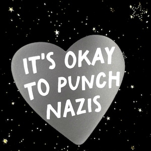 May include: A gray heart-shaped sticker with the text "IT'S OKAY TO PUNCH NAZIS" in white. The background is black with small, scattered gold stars.