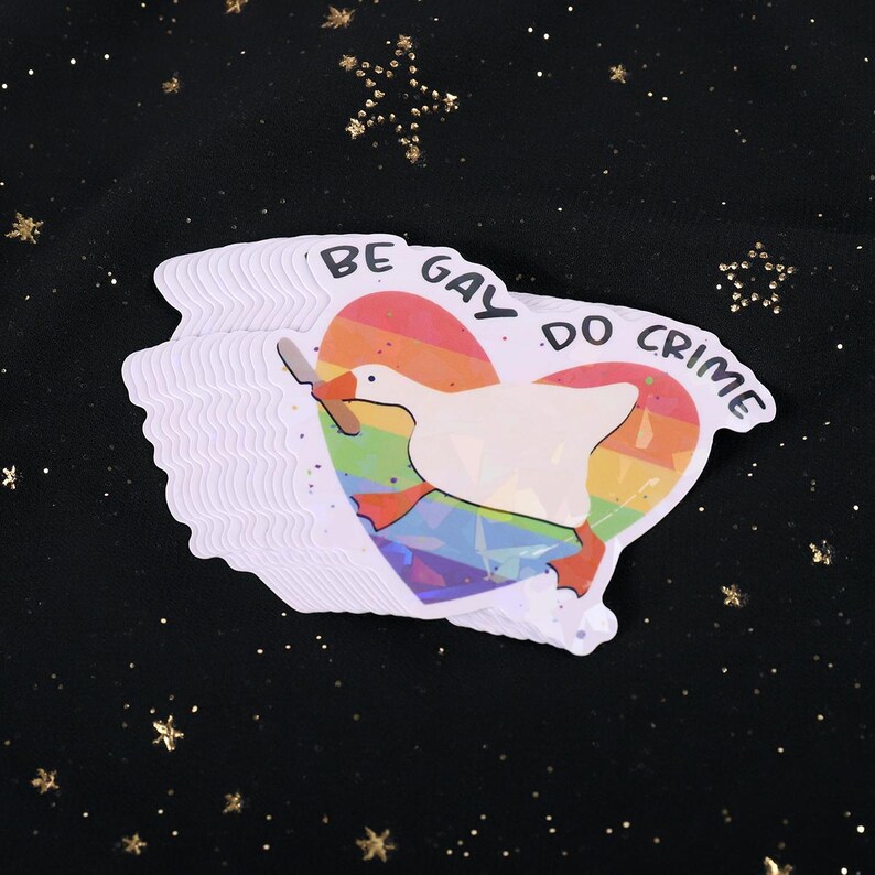 A sticker with a goose holding a knife on top of a rainbow heart, holding a knife in it's beak. The top says "Be Gay Do Crime"