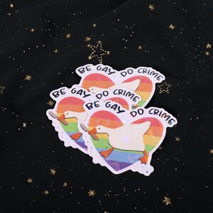 A sticker with a goose holding a knife on top of a rainbow heart, holding a knife in it's beak. The top says "Be Gay Do Crime"