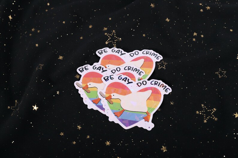 A sticker with a goose holding a knife on top of a rainbow heart, holding a knife in it's beak. The top says "Be Gay Do Crime"