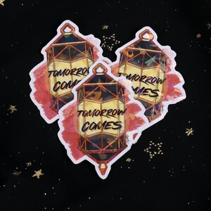 The Holographic lumina converter sticker with the text "Tomorrow Comes" on top. Behind the keychain is a red paint swipe.