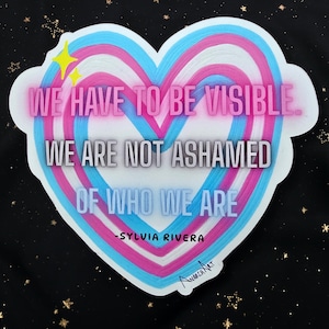 Puede incluir: Una pegatina en forma de corazón con un diseño azul, rosa y blanco. La pegatina presenta el texto "WE HAVE TO BE VISIBLE. WE ARE NOT ASHAMED OF WHO WE ARE" y el nombre "SYLVIA RIVERA". El fondo es negro con estrellas doradas.