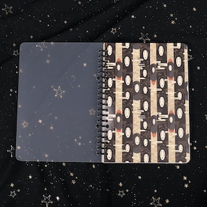 Puede incluir: Un cuaderno de espiral con una cubierta de pl&aacute;stico transparente y una cubierta estampada en blanco y negro con im&aacute;genes repetidas de Sin Cara de la pel&iacute;cula animada El viaje de Chihiro. El patr&oacute;n est&aacute; sobre un fondo beige con un sutil dise&ntilde;o de nubes.
