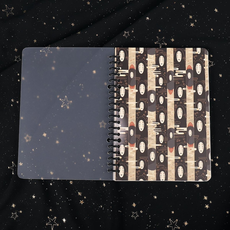 Puede incluir: Un cuaderno de espiral con una cubierta de pl&aacute;stico transparente y una cubierta estampada en blanco y negro con im&aacute;genes repetidas de Sin Cara de la pel&iacute;cula animada El viaje de Chihiro. El patr&oacute;n est&aacute; sobre un fondo beige con un sutil dise&ntilde;o de nubes.