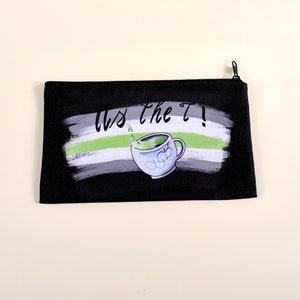 Puede incluir: Bolsa negra con cremallera con un dise&ntilde;o que presenta una taza de t&eacute; y el texto "Us the T!". El dise&ntilde;o incluye una combinaci&oacute;n de colores verde, blanco y gris, con un efecto de pincelada. La bolsa es rectangular.
