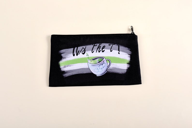 Puede incluir: Bolsa negra con cremallera con un dise&ntilde;o que presenta una taza de t&eacute; y el texto "Us the T!". El dise&ntilde;o incluye una combinaci&oacute;n de colores verde, blanco y gris, con un efecto de pincelada. La bolsa es rectangular.