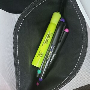 Puede incluir: Un estuche negro con cremallera que contiene un rotulador fluorescente Sharpie amarillo ne&oacute;n y dos bol&iacute;grafos Pilot Varsity. Los bol&iacute;grafos tienen tapas moradas y rosas. El estuche est&aacute; abierto, revelando su contenido.