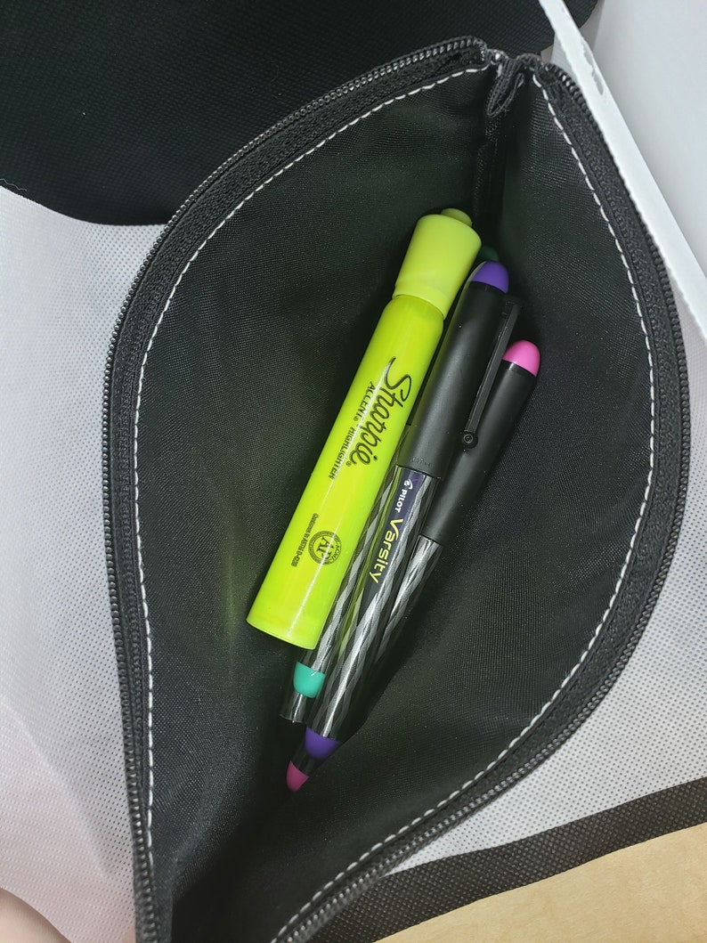 Puede incluir: Un estuche negro con cremallera que contiene un rotulador fluorescente Sharpie amarillo ne&oacute;n y dos bol&iacute;grafos Pilot Varsity. Los bol&iacute;grafos tienen tapas moradas y rosas. El estuche est&aacute; abierto, revelando su contenido.