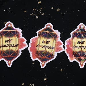 The Holographic lumina converter sticker with the text "We Continue" on top. Behind the keychain is a red paint swipe.