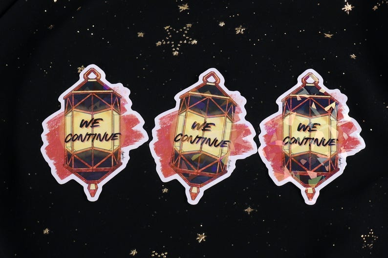 The Holographic lumina converter sticker with the text "We Continue" on top. Behind the keychain is a red paint swipe.