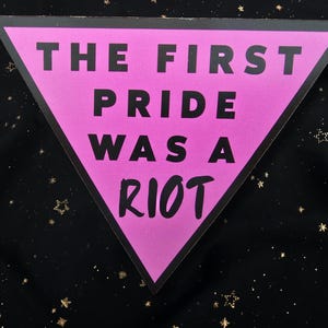 May include: A pink triangle with black text that reads "THE FIRST PRIDE WAS A RIOT." The triangle is set against a black background with gold star-shaped sparkles.