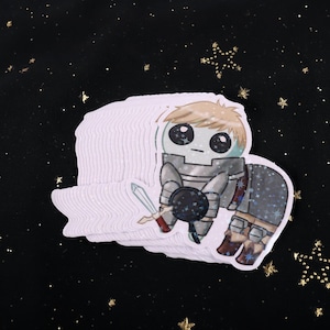 A holographic sticker Of Laios Touden  of Dungeon Meshi / Delicious in dungeon imagined as the yippee / Autism Creature. The sticker is on a black background with gold glitter.