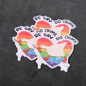 A sticker with a goose holding a knife on top of a rainbow heart, holding a knife in it's beak. The top says "Be Gay Do Crime"