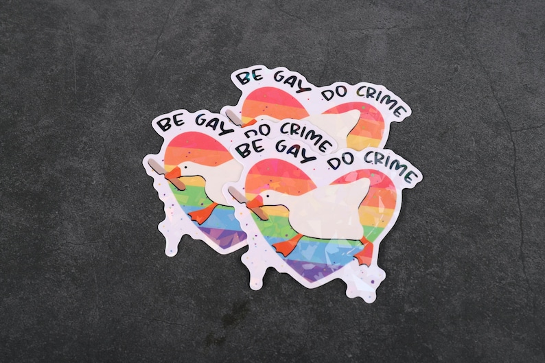 A sticker with a goose holding a knife on top of a rainbow heart, holding a knife in it's beak. The top says "Be Gay Do Crime"