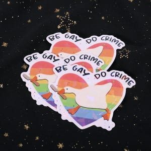 A sticker with a goose holding a knife on top of a rainbow heart, holding a knife in it's beak. The top says "Be Gay Do Crime"