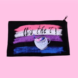 Puede incluir: Bolsa negra con cremallera con un dise&ntilde;o que presenta el texto "It's the T!" encima de un gr&aacute;fico de taza de t&eacute;. El fondo tiene pinceladas horizontales en rosa, blanco, morado y azul. La taza de t&eacute; tiene un dise&ntilde;o de galaxia.