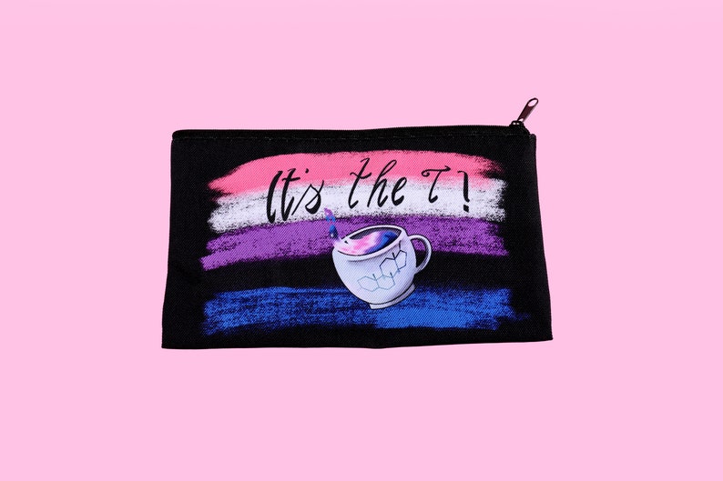 Puede incluir: Bolsa negra con cremallera con un dise&ntilde;o que presenta el texto "It's the T!" encima de un gr&aacute;fico de taza de t&eacute;. El fondo tiene pinceladas horizontales en rosa, blanco, morado y azul. La taza de t&eacute; tiene un dise&ntilde;o de galaxia.
