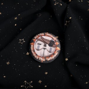 A Squishy Plush pin of Esquie from Expedition 33-  It features Esquie's head from Clair Obscur Expedition 33 featuring the text: "So the "Wheee" balances the "Whooo". He has the Expedition 33 Arm Band that Verso Gave him on his head.