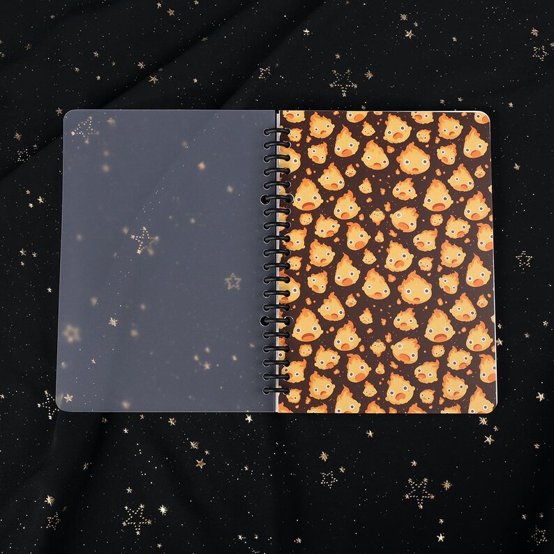 Puede incluir: Un cuaderno de espiral con una cubierta de pl&aacute;stico transparente y una cubierta colorida con un patr&oacute;n repetido de esp&iacute;ritus de fuego de dibujos animados.
