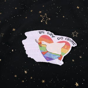 A sticker with a goose holding a knife on top of a rainbow heart, holding a knife in it's beak. The top says "Be Gay Do Crime"
