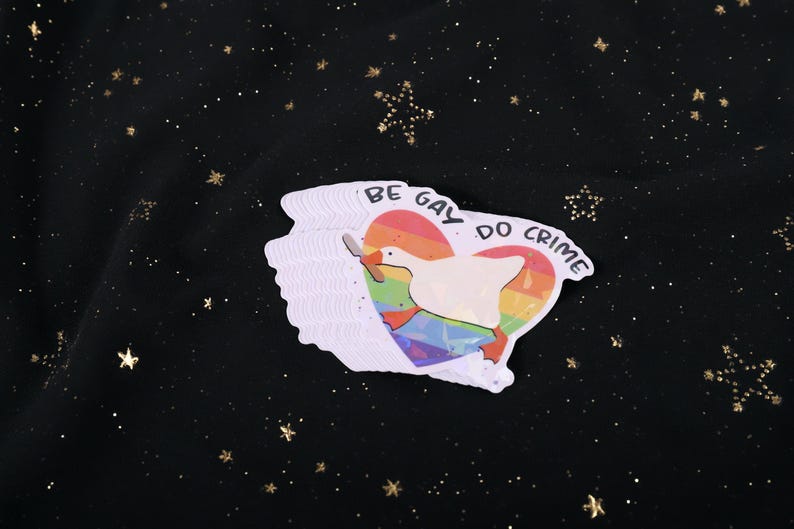A sticker with a goose holding a knife on top of a rainbow heart, holding a knife in it's beak. The top says "Be Gay Do Crime"