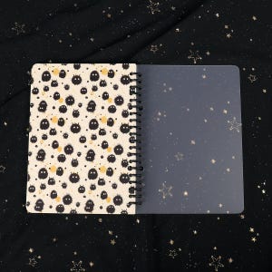 Puede incluir: Un cuaderno de espiral con un patr&oacute;n en blanco y negro de peque&ntilde;as criaturas peludas en la portada. La contraportada es transparente y tiene un sutil patr&oacute;n de estrellas.