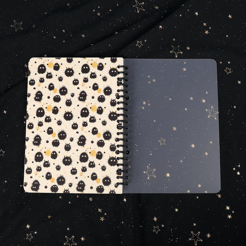Puede incluir: Un cuaderno de espiral con un patr&oacute;n en blanco y negro de peque&ntilde;as criaturas peludas en la portada. La contraportada es transparente y tiene un sutil patr&oacute;n de estrellas.