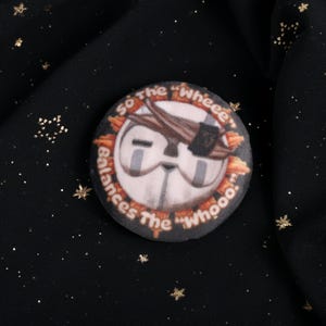 A Squishy Plush pin of Esquie from Expedition 33-  It features Esquie's head from Clair Obscur Expedition 33 featuring the text: "So the "Wheee" balances the "Whooo". He has the Expedition 33 Arm Band that Verso Gave him on his head.