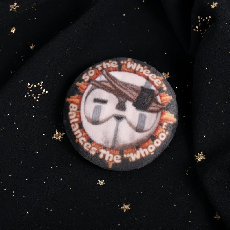A Squishy Plush pin of Esquie from Expedition 33-  It features Esquie's head from Clair Obscur Expedition 33 featuring the text: "So the "Wheee" balances the "Whooo". He has the Expedition 33 Arm Band that Verso Gave him on his head.