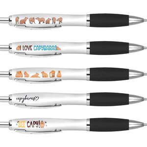 May include: Five white pens with black caps and different capybara-themed designs. The designs include capybaras in various poses, the text "I LOVE CAPYBARAS", "capybaras", and "BEE CAPY!"