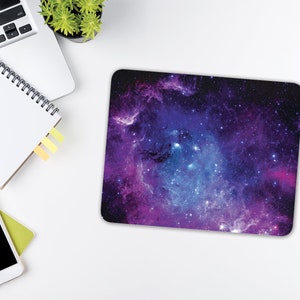 May include: A purple and blue galaxy-themed mouse mat with a nebula design. The mouse mat is on a white surface with a laptop, notebook, and smartphone.