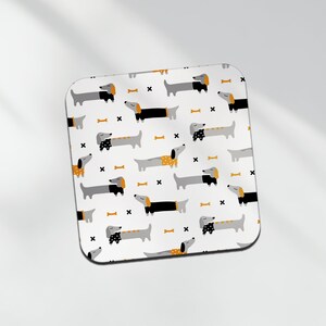 May include: A white coaster featuring a repeating pattern of black and grey dachshunds with orange accents. The dachshunds are in various poses, some lying down, some standing, and some walking. The background is white with small black crosses and orange bone shapes.