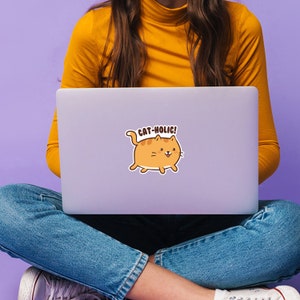 May include: A woman wearing a yellow sweater and blue jeans is sitting on a purple surface with a laptop on her lap. The laptop has a sticker of a cartoon orange cat with the text "Cat-Holic!" on it.