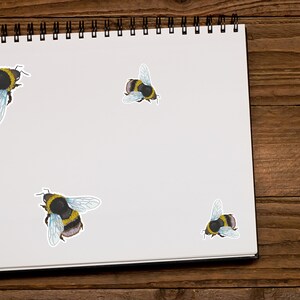 May include: A white notebook with a black spiral binding, featuring three bumble bee stickers. The notebook is on a wooden surface.