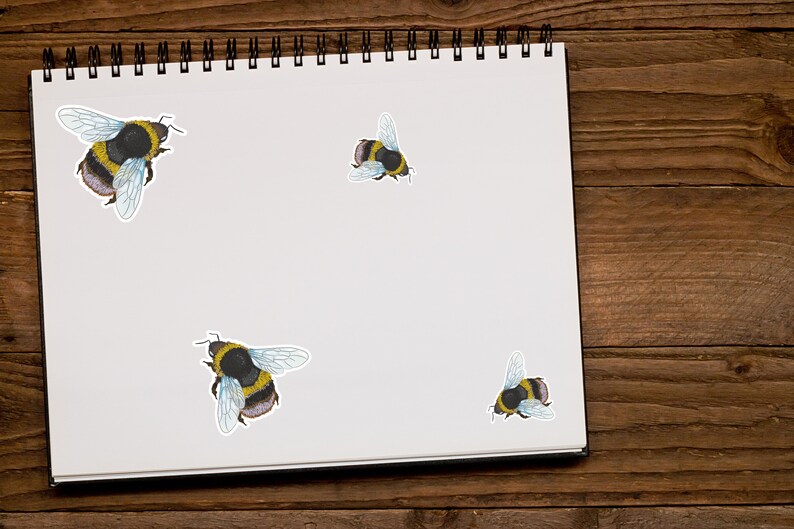 May include: A white notebook with a black spiral binding, featuring three bumble bee stickers. The notebook is on a wooden surface.
