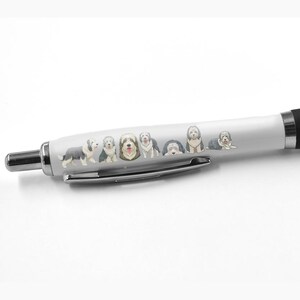 May include: A white ballpoint pen decorated with illustrations of Old English Sheepdogs. The pen has a silver metal tip and clip, and a black end. The dogs are shown in different poses, in shades of grey, white, and brown.