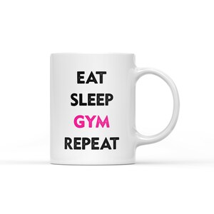 May include: White ceramic coffee mug with black text that reads "Eat Sleep Gym Repeat" with the word "Gym" in pink.
