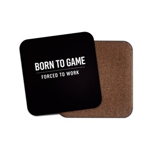 May include: Two square coasters. One is black with the text "BORN TO GAME" above "FORCED TO WORK" in white. The other coaster is brown with a cork-like texture. The coasters are designed for gaming enthusiasts.