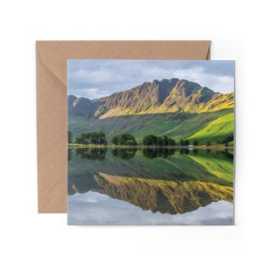 May include: A scenic photograph of a mountain range reflected in a still lake. The mountain peaks are green and brown, with a blue sky above. The lake is calm and reflects the mountains perfectly.
