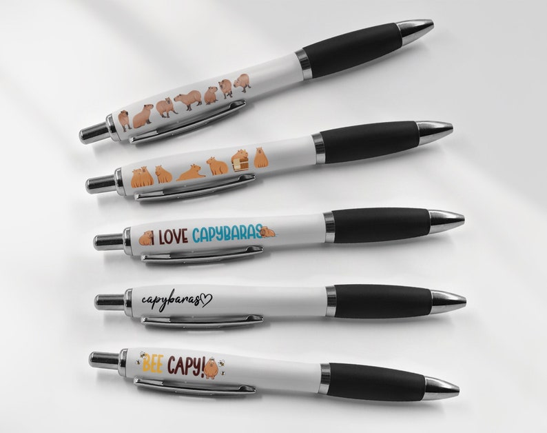 May include: Five white pens with black caps. The pens have different designs featuring capybaras, including a line of capybaras, a capybara in a house, the text "I LOVE CAPYBARAS", the text "capybaras", and the text "BEE CAPY!" with a bee and a flower.