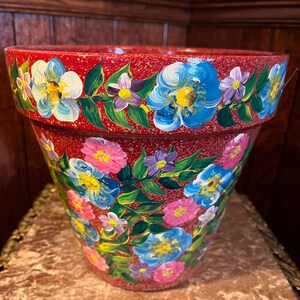 May include: A red terracotta flower pot with a hand-painted floral design. The design features blue, pink, white, and green flowers and leaves.