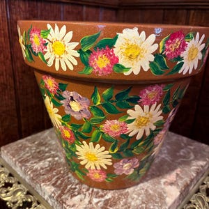 May include: A terracotta flower pot with a hand-painted design of white and pink flowers with green leaves.